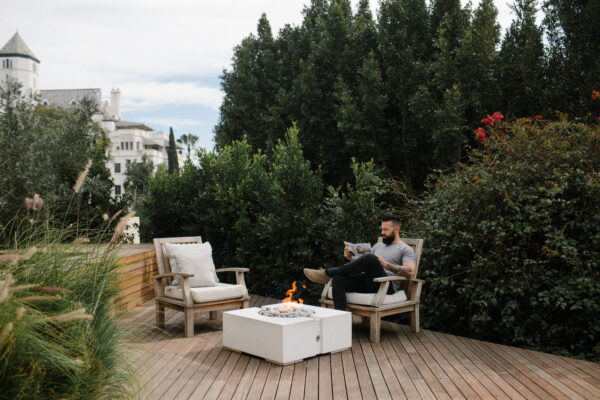 Man sitting on a wooden deck with chairs and a fire pit, surrounded by greenery.
