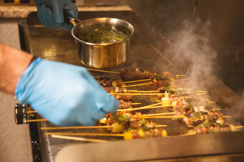 Person grilling skewered food with a pot of oil, wearing blue gloves.