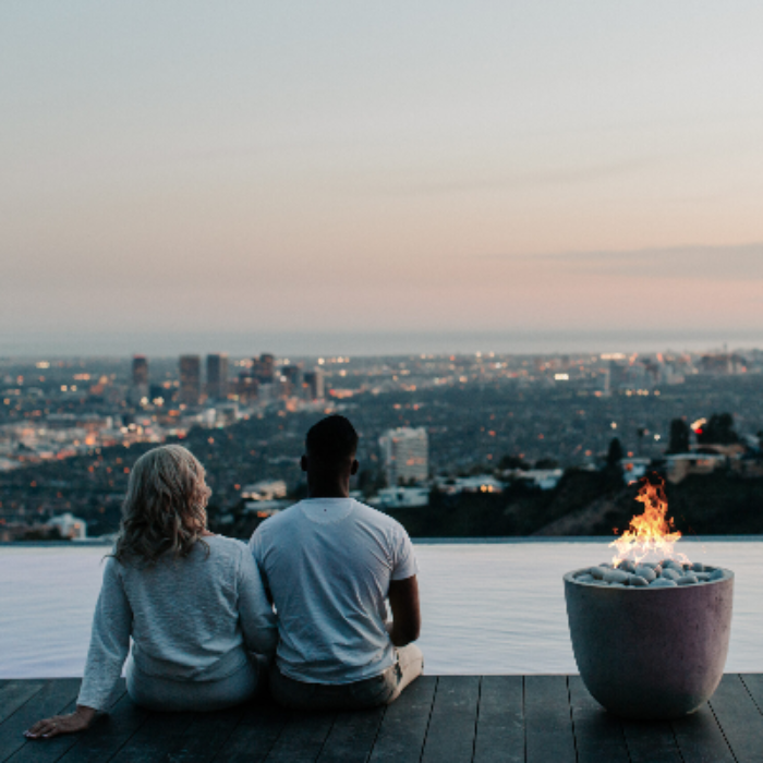 Two people sitting by a fire pit overlooking a cityscape at dusk.