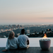 Two people sitting by a fire pit overlooking a cityscape at dusk.