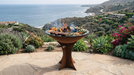 Outdoor fire pit with grill on a stone patio overlooking a scenic ocean view.